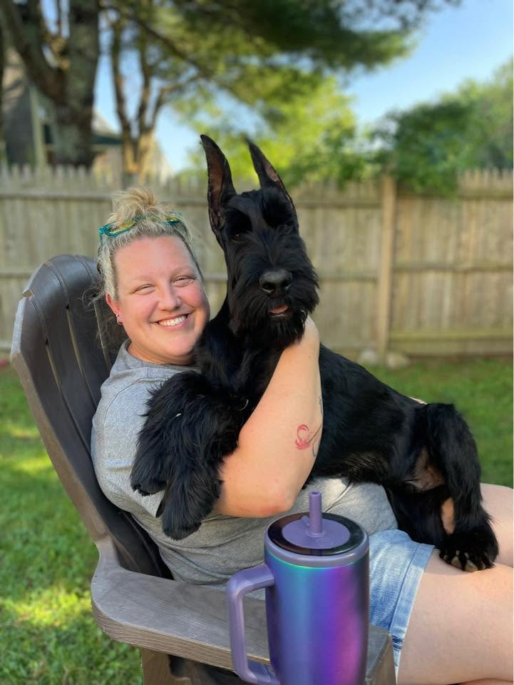 Kat smiling in a backyard chair while hugging a black Giant Schnauzer