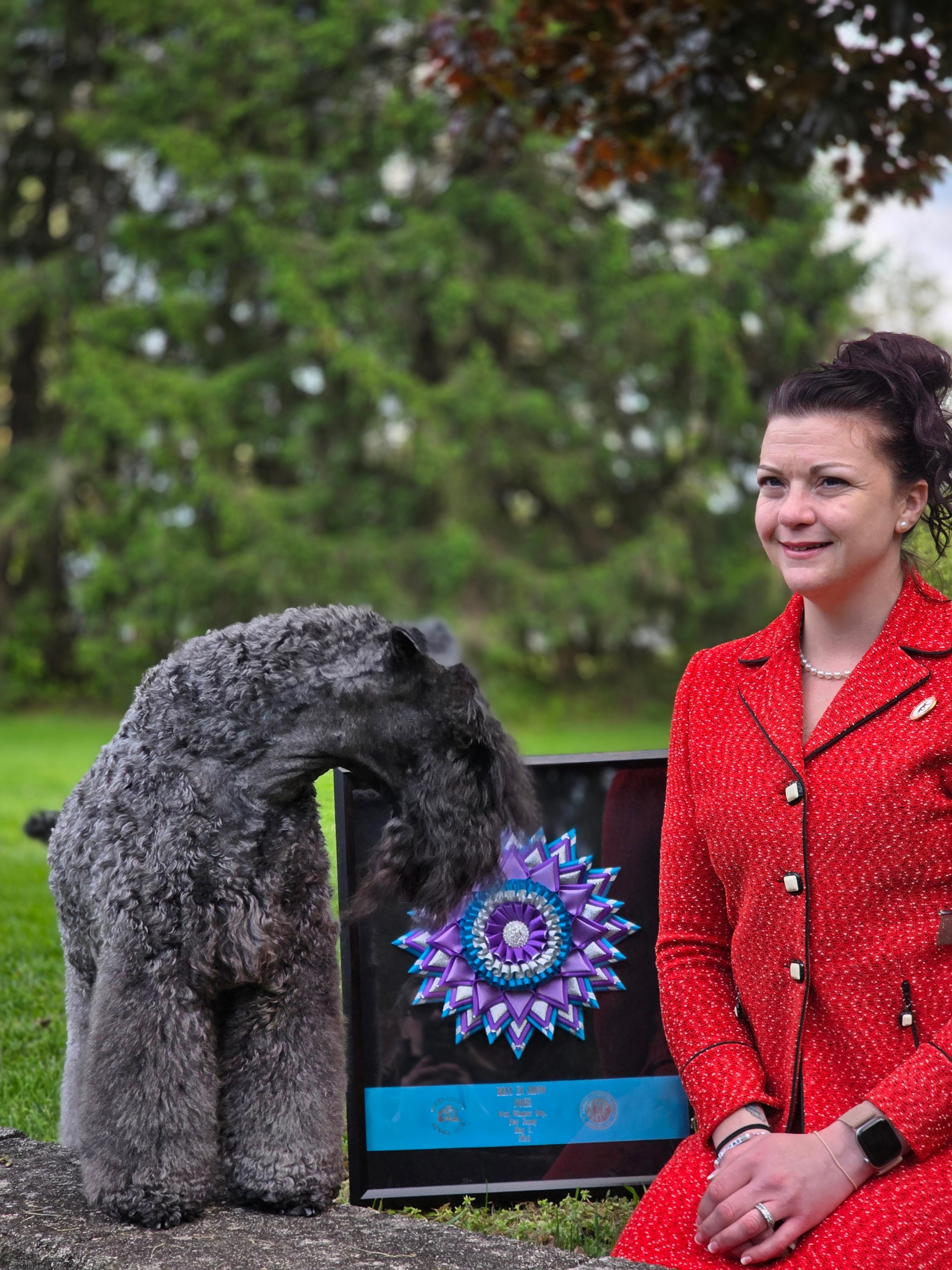 Krystal in a red jacket seated outdoors beside a groomed Kerry Blue Terrier and a framed Best in Show rosette